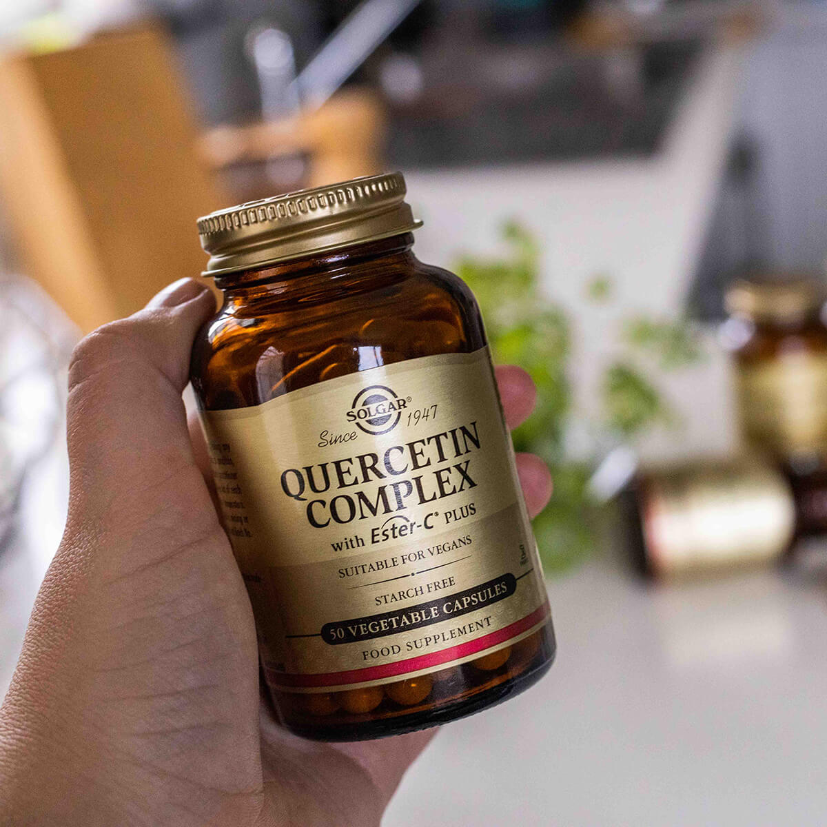 Hand holding a bottle of Solgar Quercetin Complex supplement in a kitchen setting.