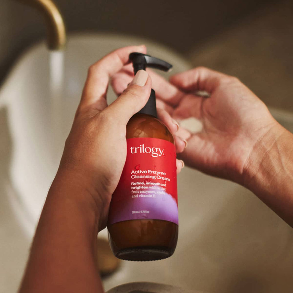 Person holding a bottle of Trilogy Active Enzyme Cleansing Cream over a sink.