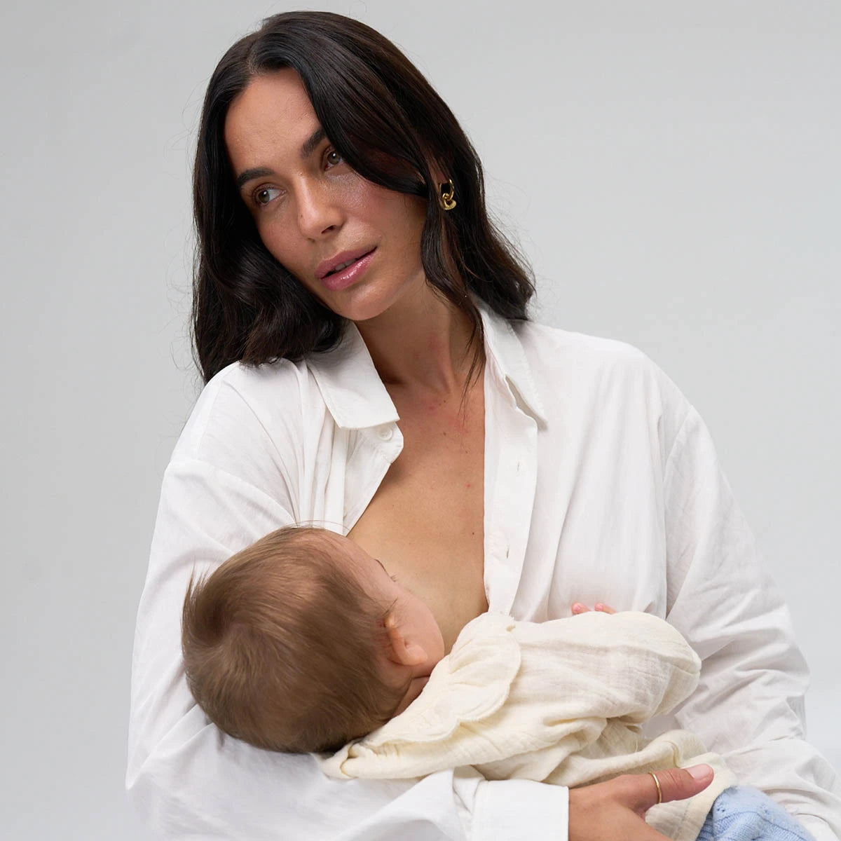 Woman standing against a grey background in a loose white shirt while breastfeeding baby.